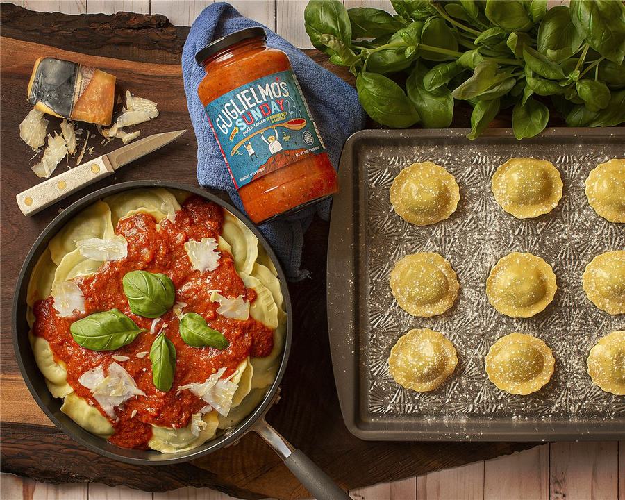 A pan of ravioli topped with Guglielmo's Sunday sauce, basil and parmesan shavings, beside the jar and a tray of fresh ravioli
