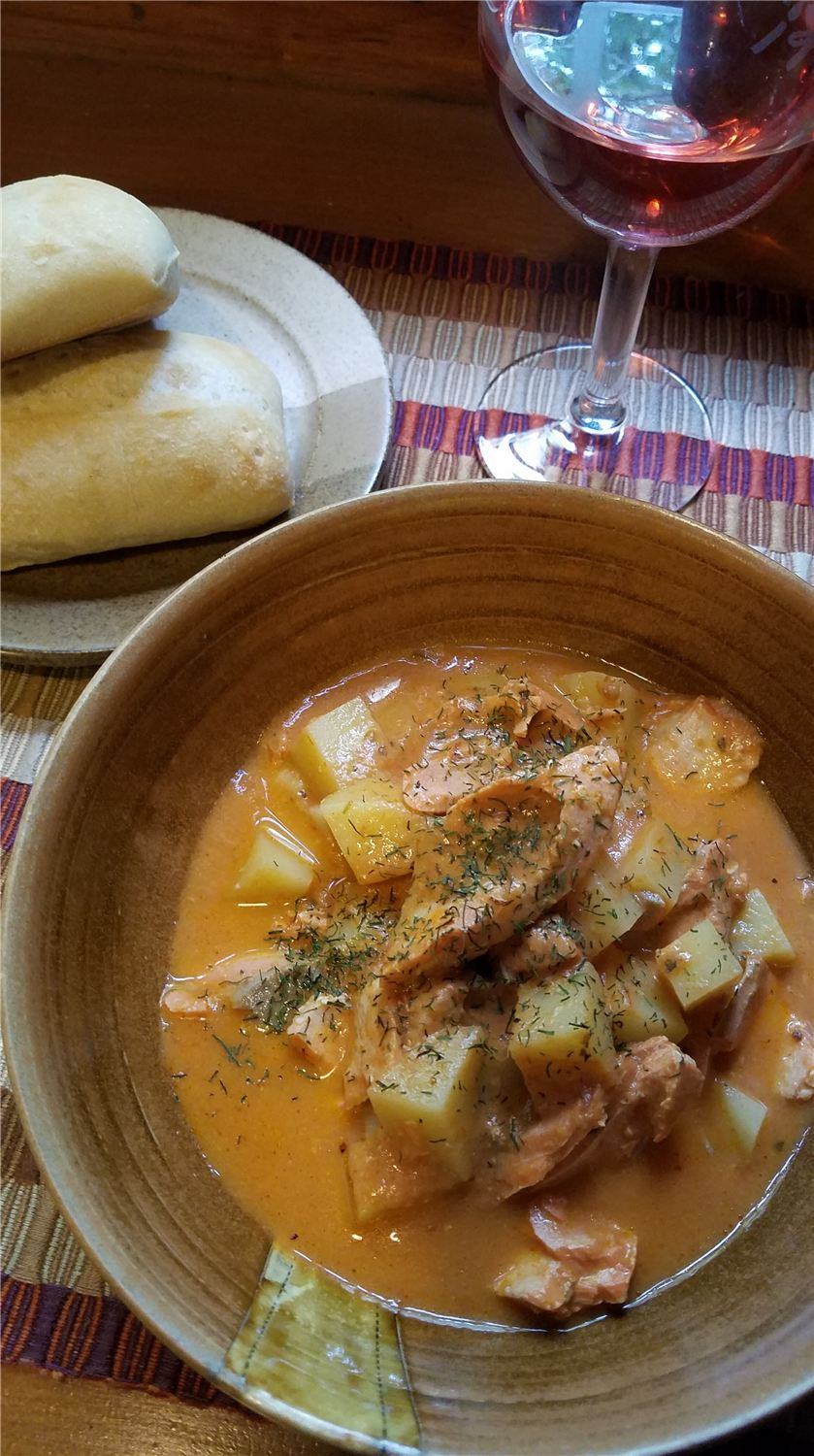 A bowl of creamy orange salmon and potato bisque with dill, beside bread rolls and a glass of rosé