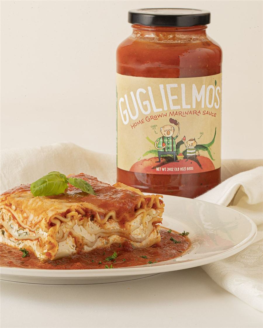 A slice of ricotta lasagna with basil on a white plate in front of a jar of Guglielmo's Home Grown Marinara Sauce