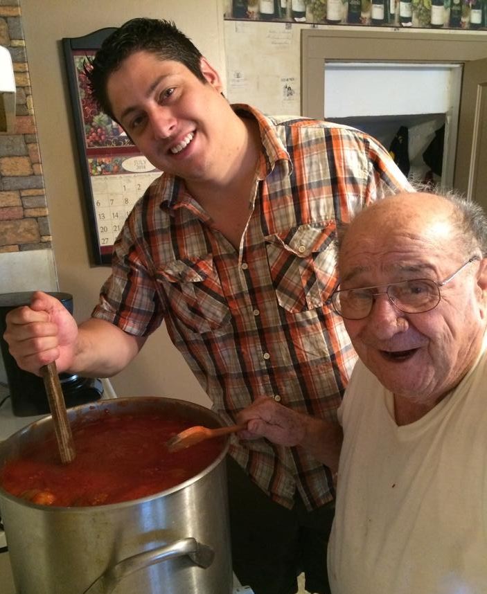 Paul and Grandpa Pete smiling beside a big steel pot of simmering tomato sauce, Paul stirring with a wooden spoon