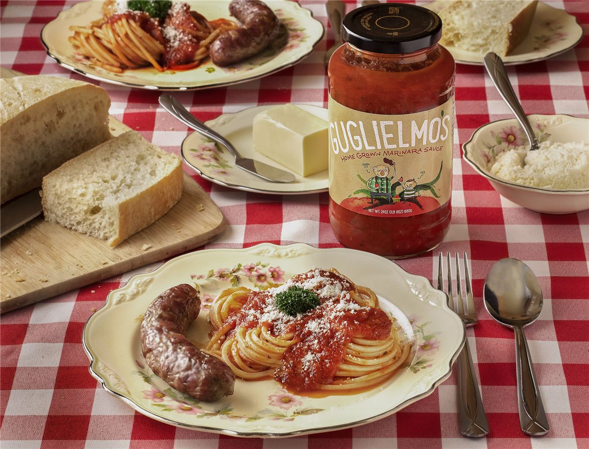 A jar of Guglielmo's Home Grown Marinara Sauce on a red gingham tablecloth beside a plate of spaghetti and sausage, with bread and parmesan